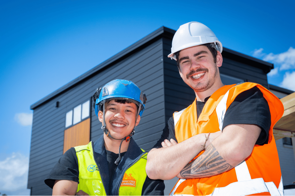 Two young trades workers wearing safety gear smile at the camera on a building site, with a modern black house in the background.