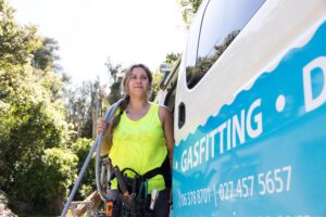 Female gasfitter in a high-vis singlet standing beside a work van, carrying equipment and wearing a tool belt.